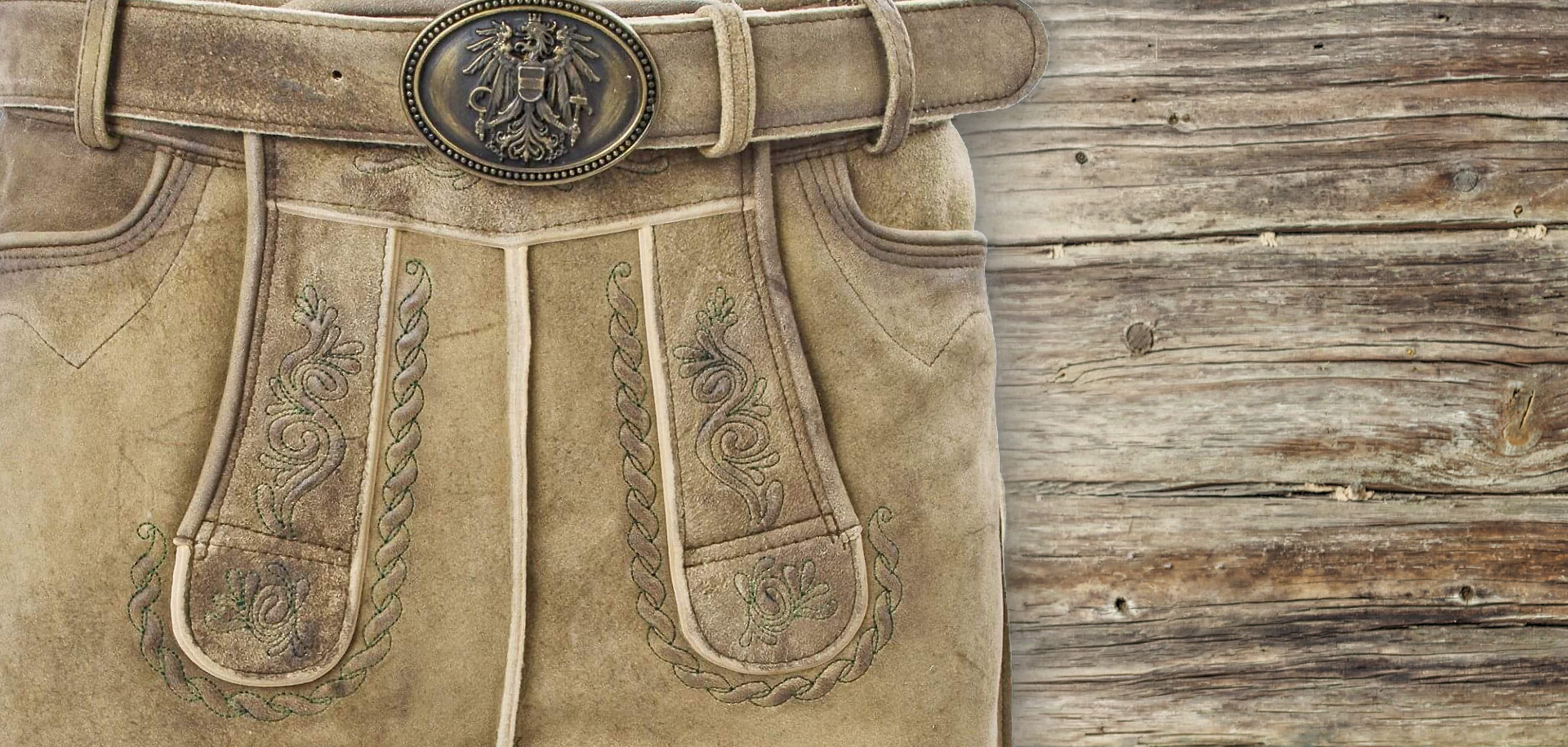 Detailansicht einer Hirschlederhose für Herren, kurze Länge, mit Fokus auf Hosenlatz und Gürtel mit Metallschnalle, rustikales Altholz im Hintergrund