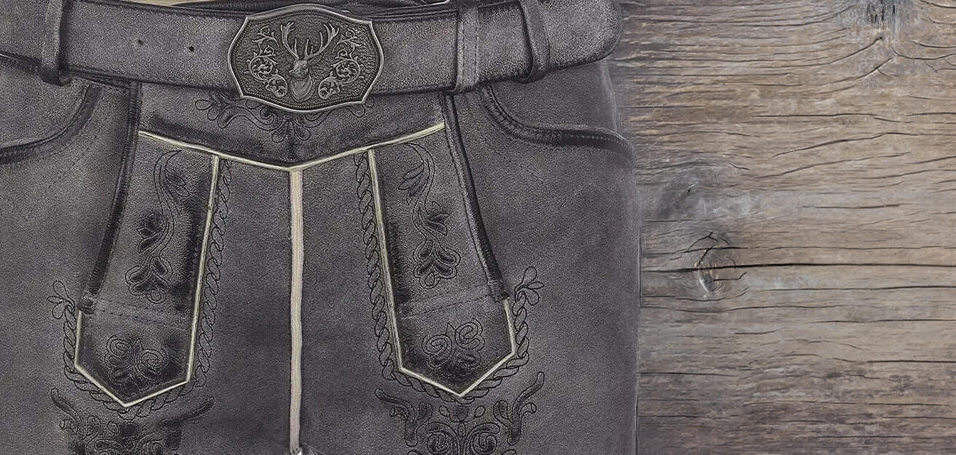 Detailansicht einer grauen Herren Lederhose, kurze Länge, mit Fokus auf Hosenlatz und Gürtel mit Metallschnalle, rustikales Holz im Hintergrund