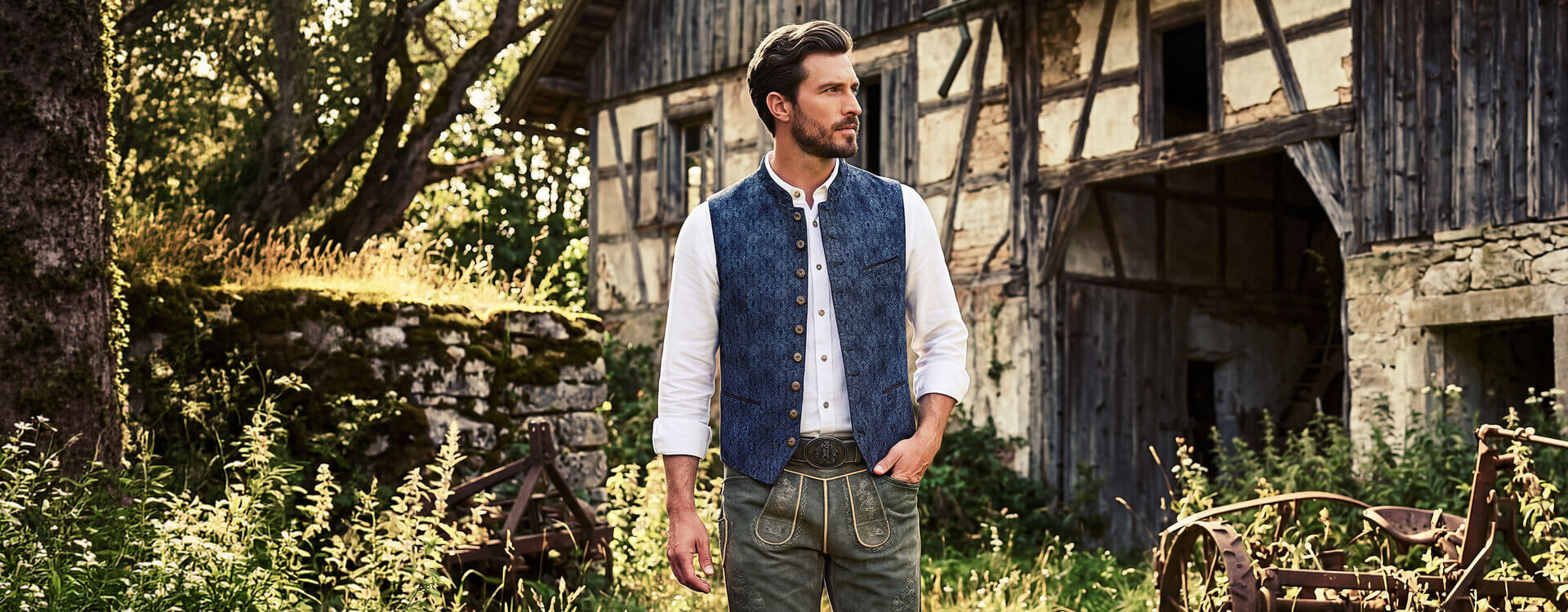 Trachtig angezogener Herr mit Lederhose und Gilet, alter Bauermhof im Hintergrund