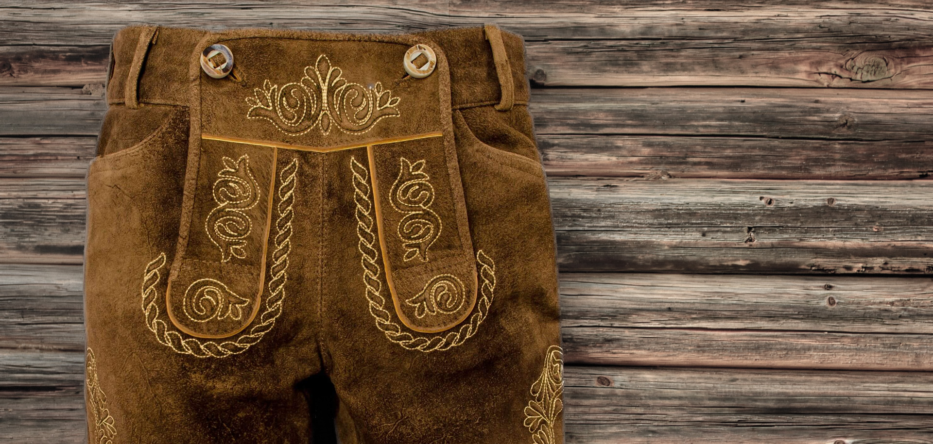 Detailansicht einer braunen Damen Lederhose, kurze Länge, mit Fokus auf Hosenlatz, rustikales Altholz im Hintergrund